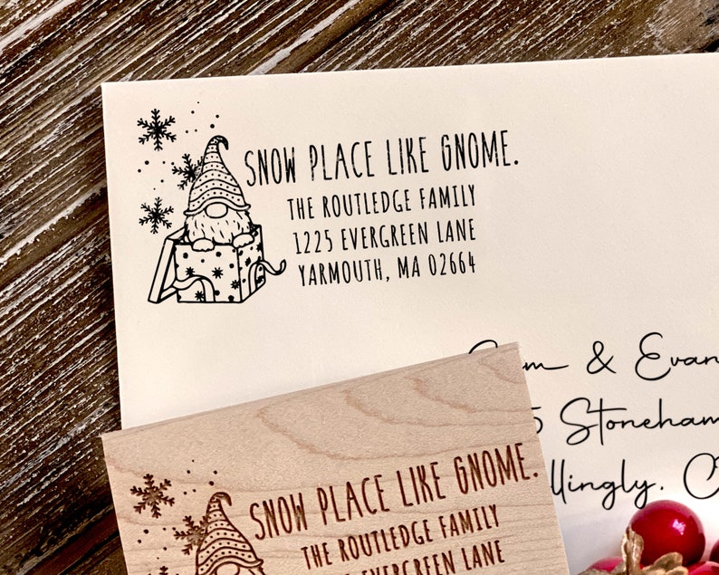 Puede incluir: Un sello de madera con un gnomo en una caja y el texto "SNOW PLACE LIKE GNOME." El texto debajo del gnomo dice "THE ROUTLEDGE FAMILY 1225 EVERGREEN LANE YARMOUTH, MA 02664".
