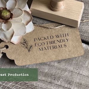 May include: A brown paper tag with a black stamp that reads "Packed with Eco Friendly Materials". The stamp also features a small leaf design. The tag is tied with twine and is sitting on a wooden surface.