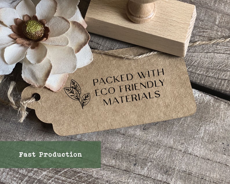 May include: A brown paper tag with a black stamp that reads "Packed with Eco Friendly Materials". The stamp also features a small leaf design. The tag is tied with twine and is sitting on a wooden surface.