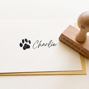 May include: A wooden rubber stamp with a round handle and a rectangular base featuring the text "stamped with love." and a small heart icon. Next to it is a white card printed with a black paw print and the name "Charlie" in script.