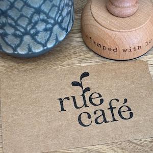 K&ouml;nnte beinhalten: Eine braune Papierkarte mit einem schwarz-wei&szlig;en Logo, auf dem "rule cafe" steht, mit einer kleinen Pflanzengrafik &uuml;ber dem Text. Die Karte liegt auf einer Holzoberfl&auml;che neben einem Holzstempel, auf dem "stamped with love" steht.