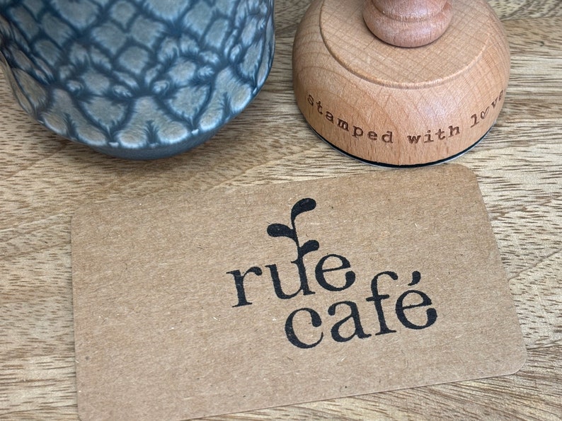 K&ouml;nnte beinhalten: Eine braune Papierkarte mit einem schwarz-wei&szlig;en Logo, auf dem "rule cafe" steht, mit einer kleinen Pflanzengrafik &uuml;ber dem Text. Die Karte liegt auf einer Holzoberfl&auml;che neben einem Holzstempel, auf dem "stamped with love" steht.