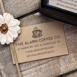 May include: A brown paper business card with the text "Five Alarm Coffee Co." and the address "67 Harris Rd. Unit B, Nashville TN". The card also includes the website "fivealarmcoffee.com" and the social media handle "@fivealarmcoffeeco".