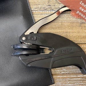 May include: A black and silver self-inking stamp with the word "trodat" printed on the side. The stamp is on a wooden surface and comes with a black leather carrying pouch.