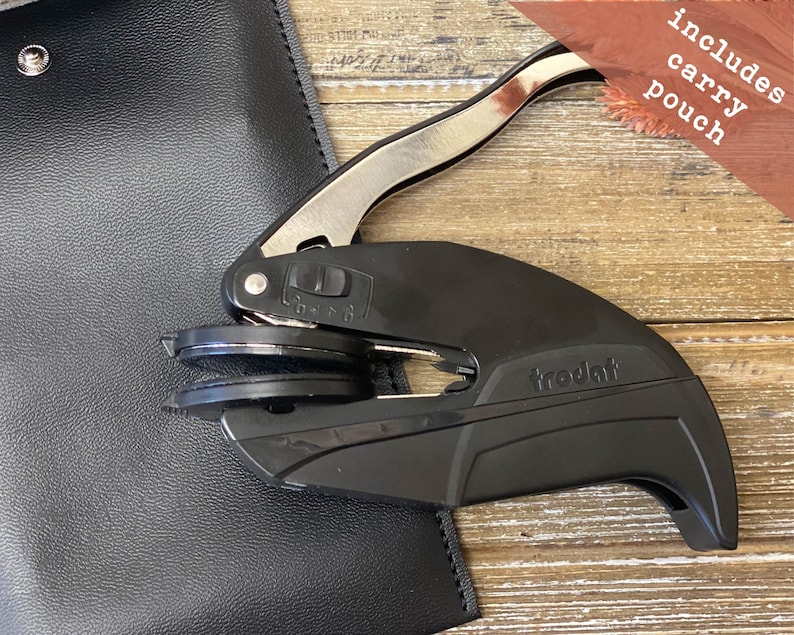 May include: A black and silver self-inking stamp with the word "trodat" printed on the side. The stamp is on a wooden surface and comes with a black leather carrying pouch.