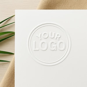 May include: A white card featuring the embossed words "YOUR LOGO" within a circular design. The card is presented against a neutral backdrop, with a touch of green foliage.