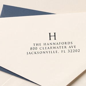 May include: A cream-colored envelope with a dark blue inner lining. The envelope is printed with the letter H, followed by the text: THE HANNAFORDS, 800 CLEARWATER AVE, JACKSONVILLE, FL 32202 in a black, serif font centered on the flap.