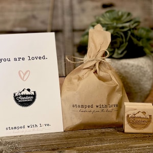 K&ouml;nnte beinhalten: Eine braune Papiert&uuml;te mit einem Schn&uuml;rverschluss, ein kleiner Holzstempel mit dem Text "Abundance Woodworks" und eine Karte mit dem Text "you are loved." und "stamped with love. handmade from the heart."