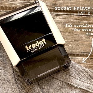 Puede incluir: Un sello Trodat Printy 4911 en blanco y negro, de 3,8 cm x 1,3 cm, dise&ntilde;ado para estampar tela. El sello est&aacute; colocado sobre una tela gris. El texto "Ink specifically for stamping fabric!" es visible.