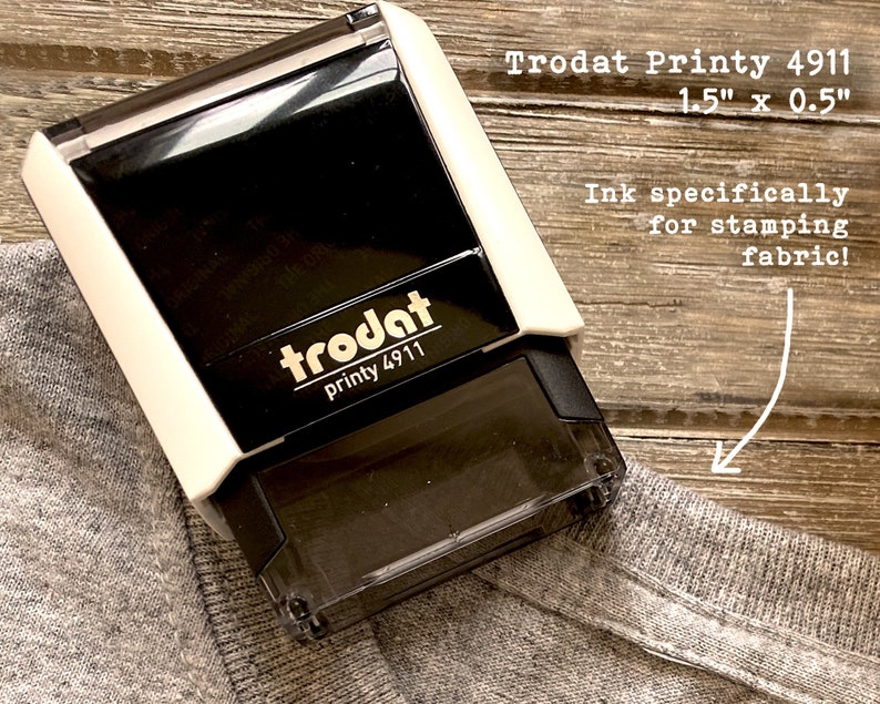 Puede incluir: Un sello Trodat Printy 4911 en blanco y negro, de 3,8 cm x 1,3 cm, dise&ntilde;ado para estampar tela. El sello est&aacute; colocado sobre una tela gris. El texto "Ink specifically for stamping fabric!" es visible.