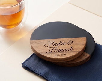 May include: Two round coasters featuring a split design with a matte black top half and a natural wood grain bottom half. The wood section is engraved with the text Andre & Hannah and EST. 2026 in a script and sans-serif font combination.