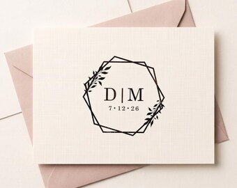 May include: A cream-colored rectangular card featuring a black geometric frame with leaf accents. The center displays the letters D | M in a serif font, with the date 7 · 12 · 26 printed below. The card is positioned over a light pink envelope.