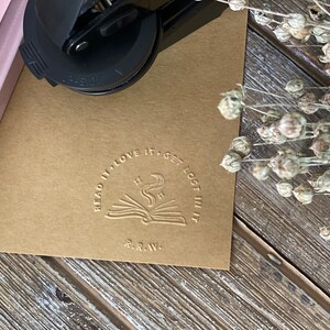 May include: A brown paper card with a round, embossed stamp that reads "Read it, love it, get lost in it." The stamp features a stylized image of an open book with a rising plume of smoke and stars above it. The text "R.R.W." is below the image.