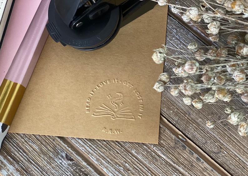 May include: A brown paper card with a round, embossed stamp that reads "Read it, love it, get lost in it." The stamp features a stylized image of an open book with a rising plume of smoke and stars above it. The text "R.R.W." is below the image.