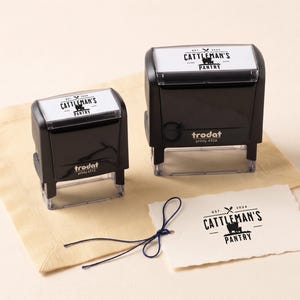 Op de afbeelding: Twee zwarte Trodat Printy zelfinktende stempels, modellen 4913 en 4926, voorzien van de tekst CATTLEMAN'S PANTRY en een koe-silhouet. Een wit rechthoekig label met hetzelfde gestempelde logo en een donkerblauw touwtje ligt op de voorgrond.