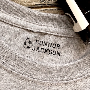 K&ouml;nnte beinhalten: Ein graues T-Shirt mit einem schwarzen Fu&szlig;ball und dem Namen "CONNOR JACKSON" auf der Vorderseite.