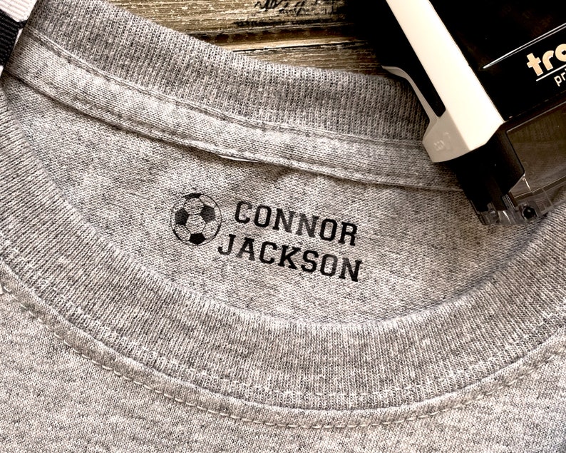 K&ouml;nnte beinhalten: Ein graues T-Shirt mit einem schwarzen Fu&szlig;ball und dem Namen "CONNOR JACKSON" auf der Vorderseite.