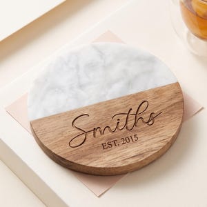 May include: A round coaster featuring a split design with white marble on the top half and natural brown wood on the bottom half. The wood section is engraved with the text "Smiths" in a script font and "EST. 2015" in a block font.