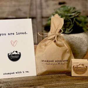 May include: A brown paper bag with a string tie, a small wooden stamp with the text "Abundance Woodworks" and a card with the text "you are loved." and "stamped with love. -handmade from the heart-"