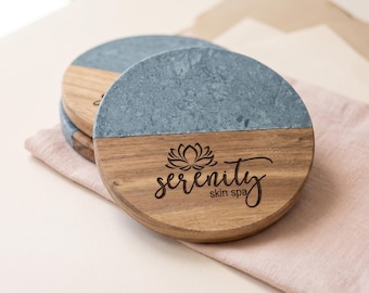 Puede incluir: Una pila de posavasos redondos con un diseño dividido de piedra gris y madera marrón natural. La parte de madera está grabada con un icono de flor de loto y el texto Serenity skin spa en una fuente negra cursiva y sans-serif.