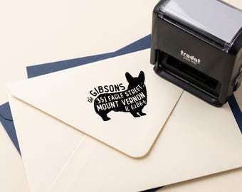 May include: A black Trodat Printy 4926 self-inking stamp next to a cream envelope. The stamp has imprinted a black silhouette of a dog containing the text: THE GIBSONS, 351 EAGLE STREET, MOUNT VERNON, IL 62864, clearly visible on the envelope flap.