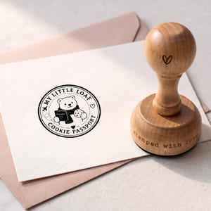 May include: A wooden rubber stamp with a rounded handle featuring a heart icon and the text stamped with love. The stamp creates a circular black ink impression of a bear holding a passport and a cookie with the text MY LITTLE LOAF COOKIE PASSPORT.