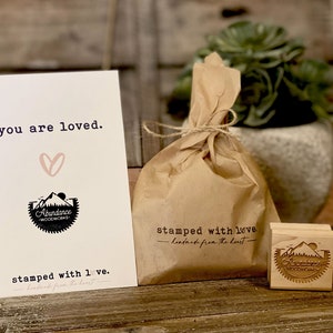 Puede incluir: Una tarjeta con el texto "you are loved" y un coraz&oacute;n, una bolsa de papel marr&oacute;n atada con hilo y un sello de madera. El sello y la tarjeta presentan el logotipo "Abundance Woodworks".