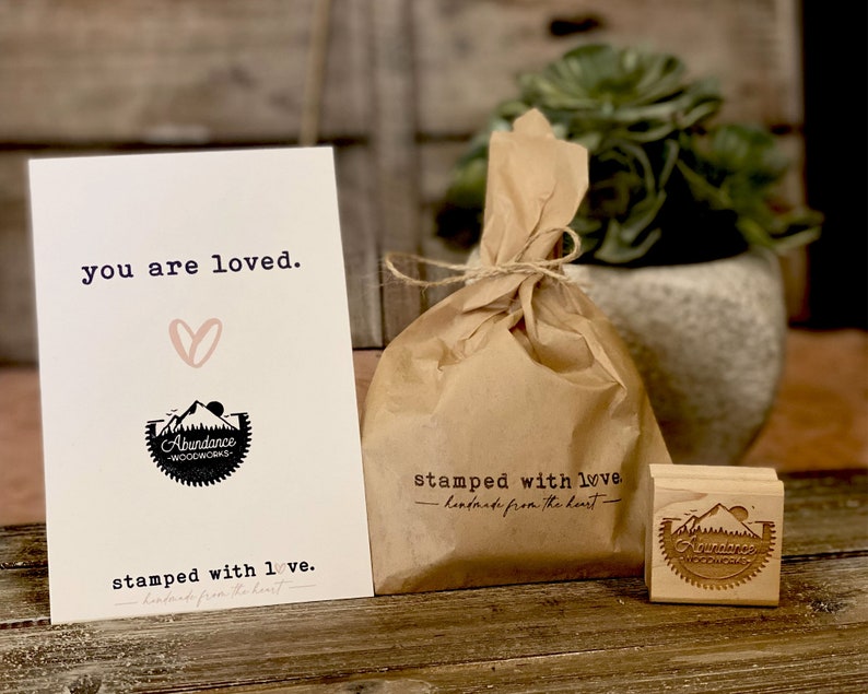 Puede incluir: Una tarjeta con el texto "you are loved" y un coraz&oacute;n, una bolsa de papel marr&oacute;n atada con hilo y un sello de madera. El sello y la tarjeta presentan el logotipo "Abundance Woodworks".