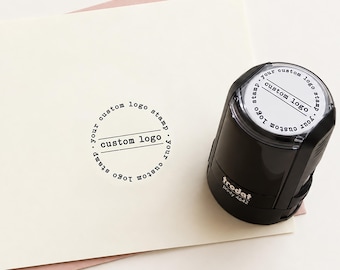 May include: A black Trodat Printy 4642 self-inking stamp with a circular impression area. The stamp features the text 'custom logo' in the centre with 'your custom logo stamp' written in a circle around the perimeter on a white paper surface.