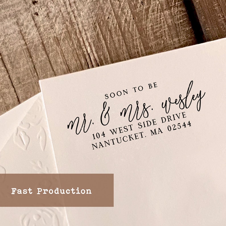 May include: A white card with black text that reads "SOON TO BE Mr. & Mrs. wesly 104 WEST SIDE DRIVE NANTUCKET. MA 02544". The text is in a cursive font. The card is on a wooden surface.