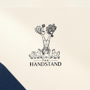 May include: A square off-white greeting card featuring a black ink illustration of a person in a handstand wearing a hat and overalls. The person balances vegetables and flowers on their feet, surrounded by three chickens. Text reads THE HANDSTAND.