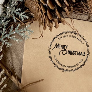 Peut inclure: Une carte en papier brun avec un tampon d'encre noire qui indique "The McGregor Family Merry Christmas 54 Outlander Dr. Portland, OR 97035" dans un design de couronne.