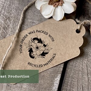 May include: A brown paper tag with a black stamp that reads "Your order was packed with recycled materials". The stamp features a stylized recycling symbol with flowers and leaves. The tag is tied with twine.