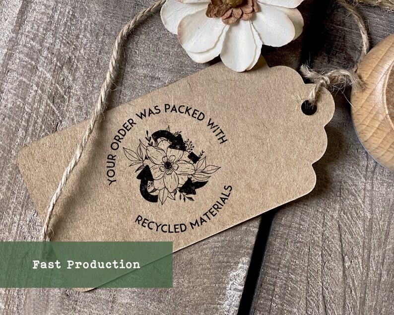 May include: A brown paper tag with a black stamp that reads "Your order was packed with recycled materials". The stamp features a stylized recycling symbol with flowers and leaves. The tag is tied with twine.