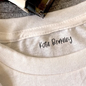 Puede incluir: Primer plano de un cuello de camiseta blanca con el nombre "Katie Berkeley" impreso en escritura negra. Una camiseta gris y una grapadora son visibles en el fondo. La imagen se centra en la textura de la camiseta y el texto.