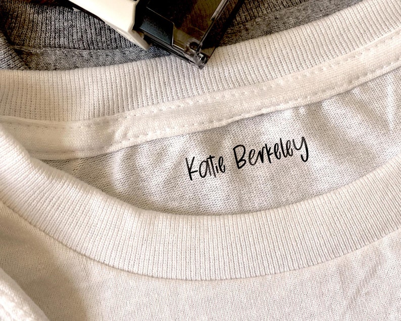 Puede incluir: Primer plano de un cuello de camiseta blanca con el nombre "Katie Berkeley" impreso en escritura negra. Una camiseta gris y una grapadora son visibles en el fondo. La imagen se centra en la textura de la camiseta y el texto.