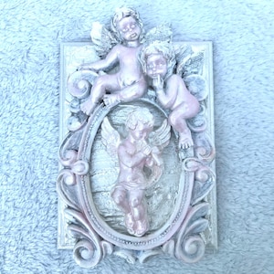 Könnte beinhalten: Rechteckige Wandplakette mit drei geflügelten Cherubim. Die Cherubim sind in einem hellrosa Farbton gehalten, vor einem weißen und grauen verzierten Rahmen. Die Plakette hat eine strukturierte Oberfläche und ist wahrscheinlich aus Kunstharz.