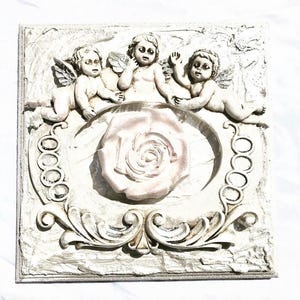 Puede incluir: Escultura en relieve cuadrada, blanquecina, con tres figuras de querubines alados, rodeando un diseño central de rosa. La escultura tiene detalles clásicos ornamentados y probablemente está hecha de yeso o material similar.