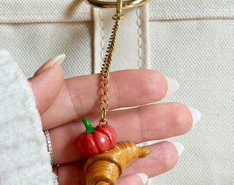 Op de afbeelding: Een goudkleurige kettinghanger met een miniatuur croissant en een rode pompoen. De hanger is bevestigd aan een gouden ring. De hanger wordt voor een witte draagtas gehouden.
