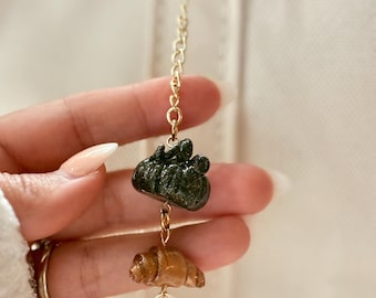May include: A gold-colored keychain with a charm featuring a dark green pumpkin, a brown croissant, and a gray pearl. The charm is suspended from a gold chain and attached to a gold ring.