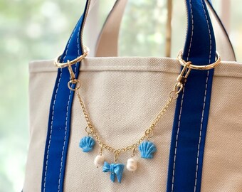 May include: A cream canvas tote bag with blue trim and straps. A gold chain with blue seashell charms, a blue bow, and white pearl charms hangs from the bag's handles. The bag has a nautical theme.