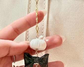 Puede incluir: Un llavero dorado con una calabaza blanca, un lazo negro con purpurina y una perla blanca. El dije cuelga de un anillo y una cadena dorados, creando un accesorio decorativo.