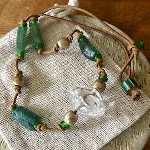 May include: A necklace featuring a tan leather cord strung with cylindrical green glass beads, round metallic beads, and a central clear, textured glass bead. The cord is knotted between the beads, and a striped green and white bead is near the end.