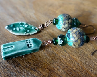 May include: A pair of green ceramic earrings with a house and leaf charm on one side and a green and brown patterned bead on the other.