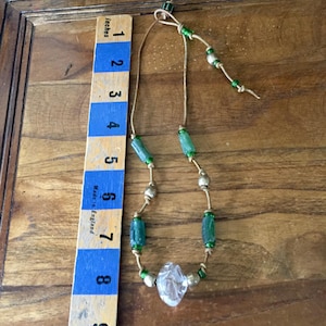 May include: A necklace featuring a tan leather cord strung with green cylindrical beads, metallic spherical beads, and a single clear, faceted central bead. A wooden ruler placed alongside shows the necklace length is approximately 8 inches.