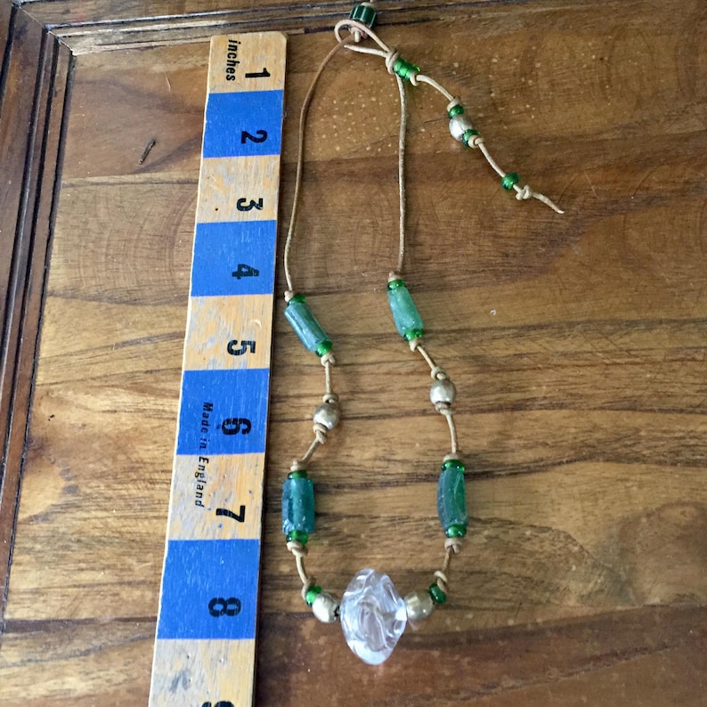 May include: A necklace featuring a tan leather cord strung with green cylindrical beads, metallic spherical beads, and a single clear, faceted central bead. A wooden ruler placed alongside shows the necklace length is approximately 8 inches.