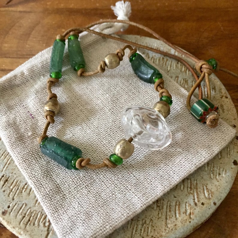 May include: A necklace featuring a tan leather cord strung with cylindrical green glass beads, round metallic beads, a clear faceted glass center bead, and a striped green and white glass bead at the end, all secured with knotted leather cord details.