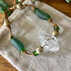 May include: A beaded necklace featuring a central clear faceted glass bead, interspersed with matte gold-toned spherical beads, cylindrical sea-glass green beads, and small translucent green glass spacer beads on a tan leather cord.