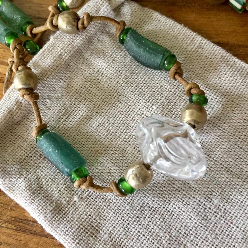May include: A beaded necklace featuring a central clear faceted glass bead, interspersed with matte gold-toned spherical beads, cylindrical sea-glass green beads, and small translucent green glass spacer beads on a tan leather cord.
