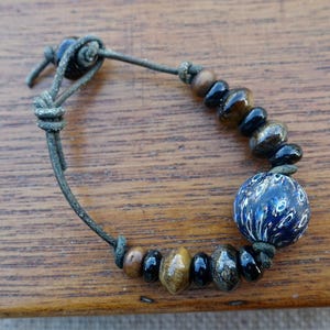 May include: A bracelet featuring a dark leather cord with a sliding knot closure. The cord is strung with a variety of beads, including small black, brown, and metallic-toned beads, and one large, round, dark blue bead with white speckled patterns.